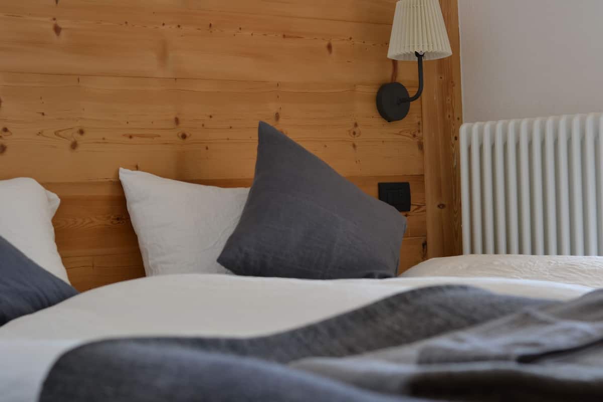 A neatly made bed with white and gray bedding, a wooden headboard, a wall-mounted lamp, and a radiator.
