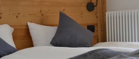 Una cama bien hecha con ropa de cama blanca y gris, un cabecero de madera, una lámpara de pared y un radiador.