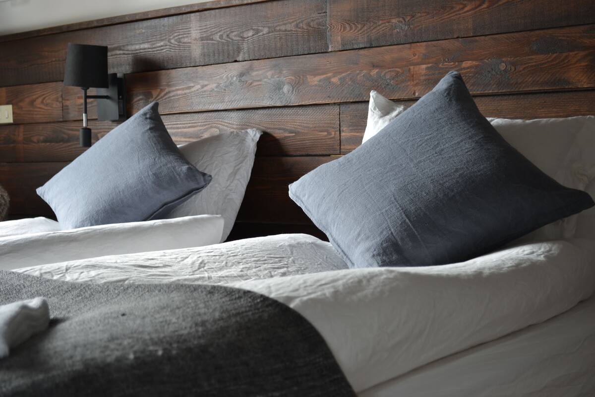 A bed with white linens and two grey pillows, set against a wooden headboard with a black wall-mounted lamp.