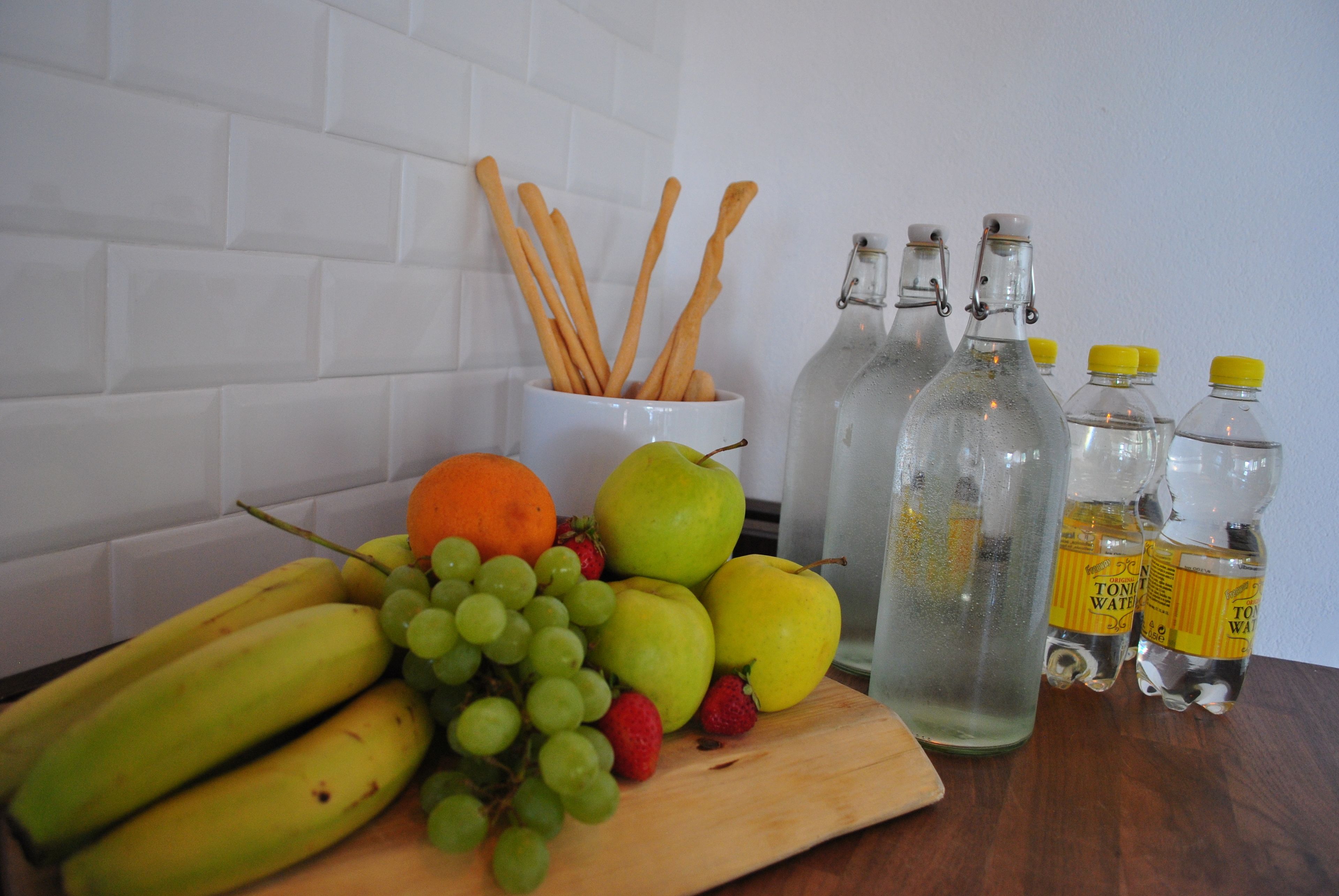 A wooden cutting board with fruits, a white mug with wooden utensils, and three glass bottles with yellow caps.