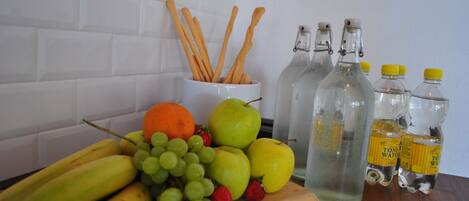 A wooden cutting board with fruits, a white mug with wooden utensils, and three glass bottles with yellow caps.