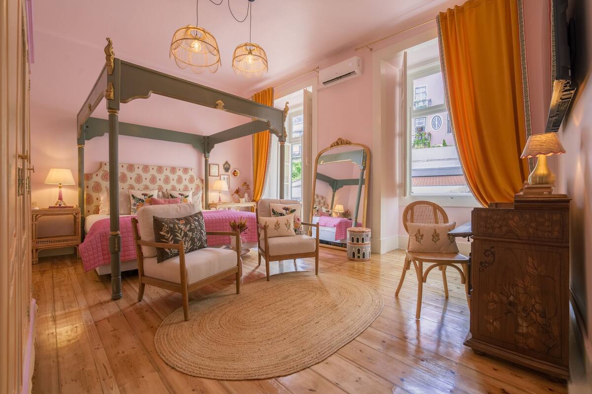 A four-poster bed with a canopy, a sitting area with a chair and a small table, a wooden cabinet, and a large mirror.