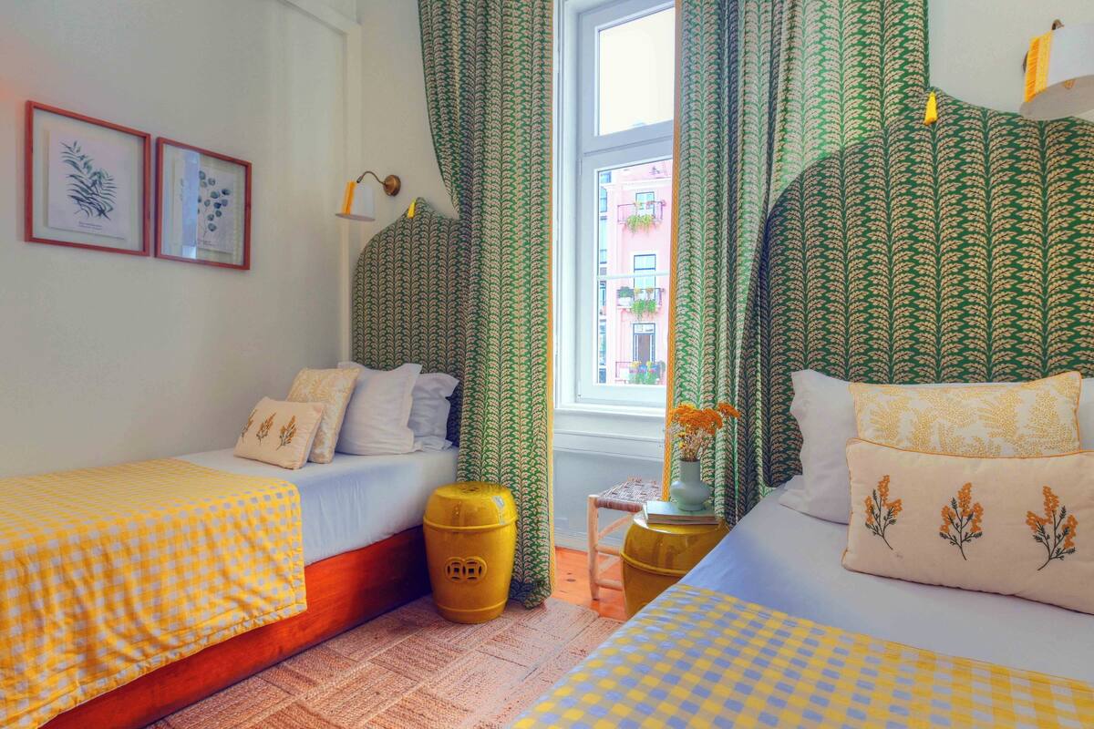 A room with two beds, green patterned curtains, and a window with a view of buildings.