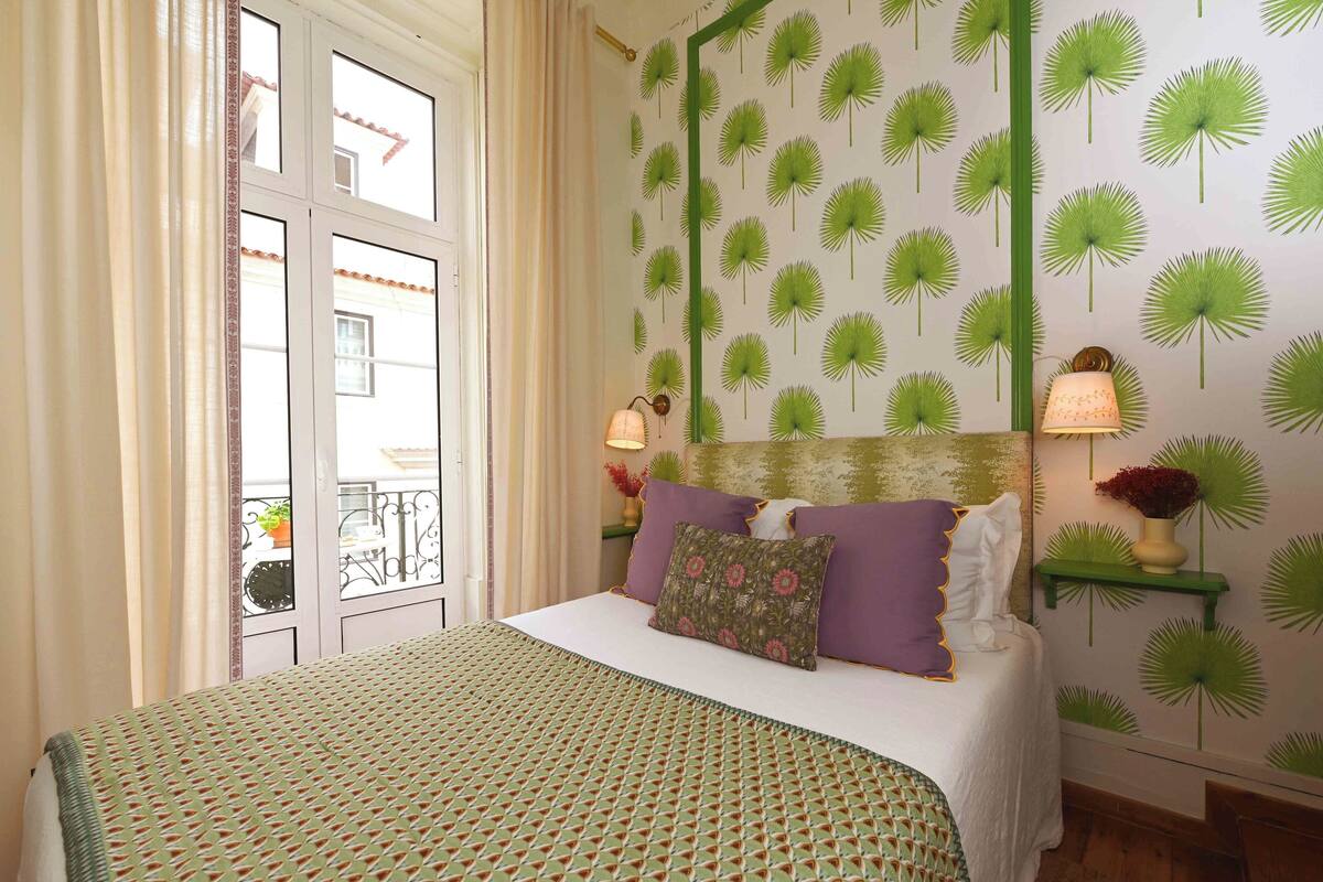 A bedroom with a bed, a window with curtains, a wall with a green palm leaf pattern, and a bedside table with a lamp.