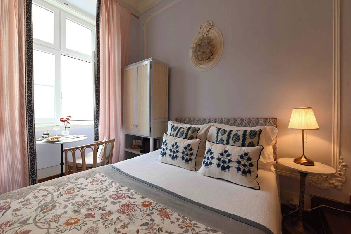 A hotel room with a bed, a bedside table with a lamp, a chair, a small table with a vase, and a wardrobe.