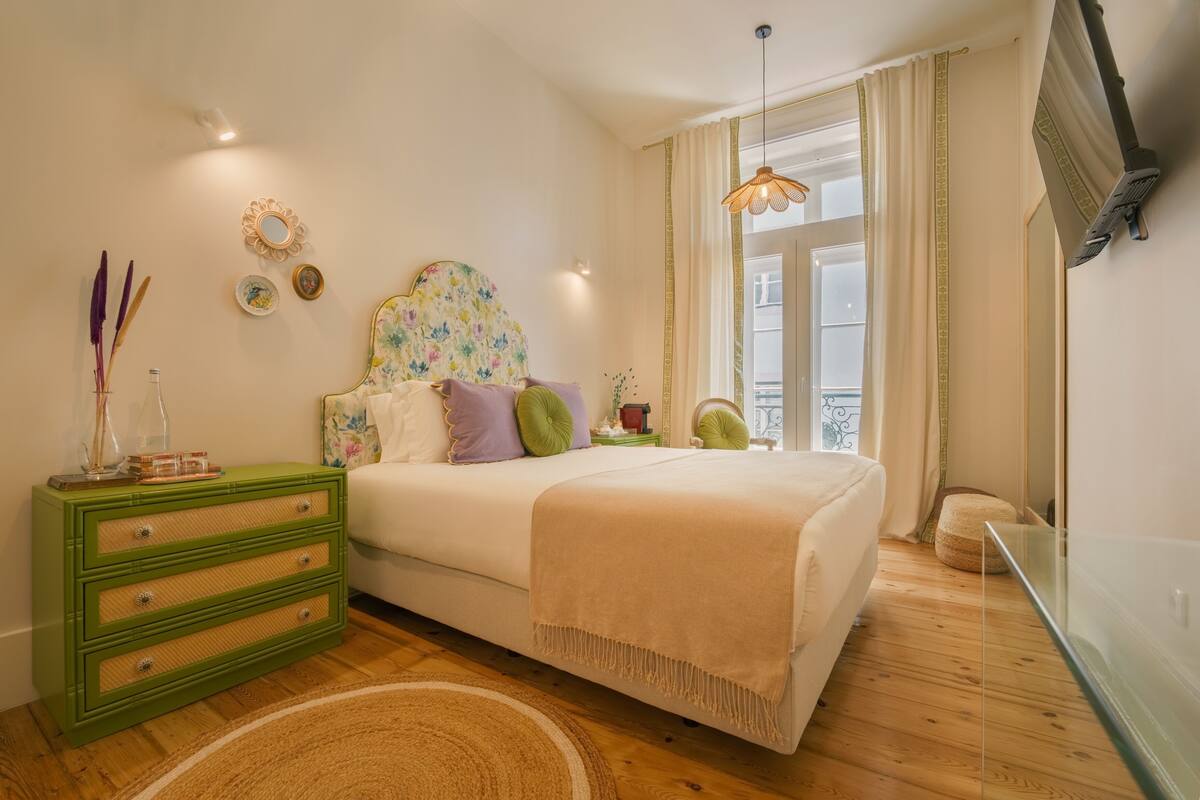 A bedroom with a bed, green dresser, decorative mirror, and a window with curtains.