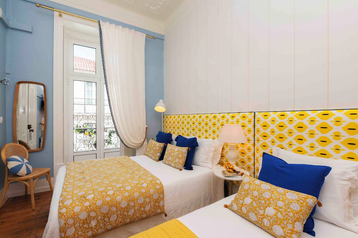 A hotel room with two beds, a window with curtains, a mirror, and a patterned headboard.