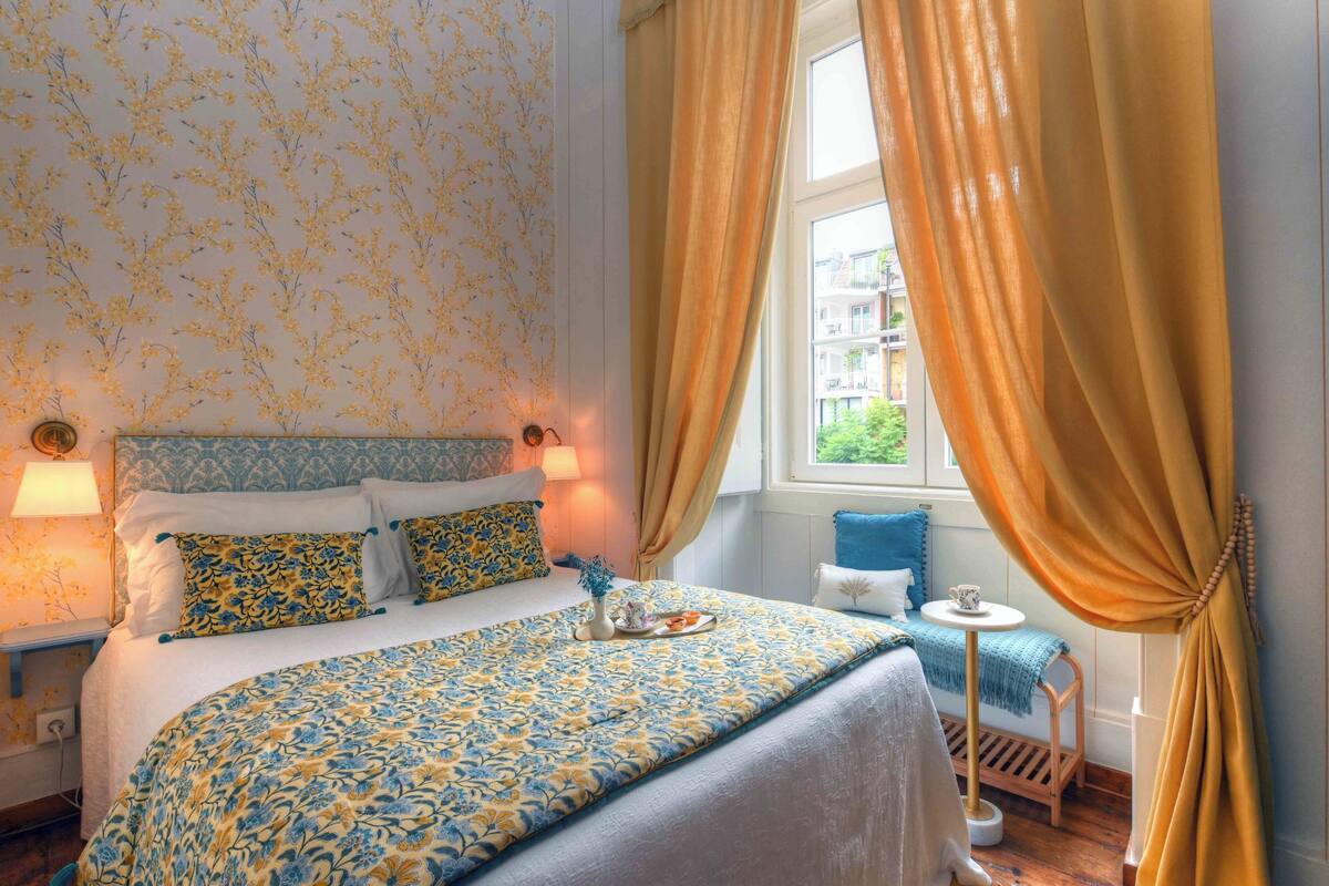 A bedroom with a floral-patterned bedspread, a bedside table with a lamp, and a view of the outside through a window with curtains.