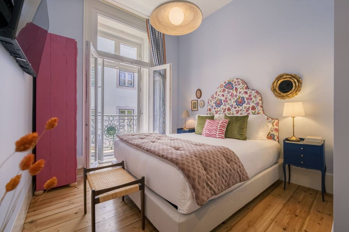 A bedroom with a bed, bedside table, lamp, and a view of the outside.