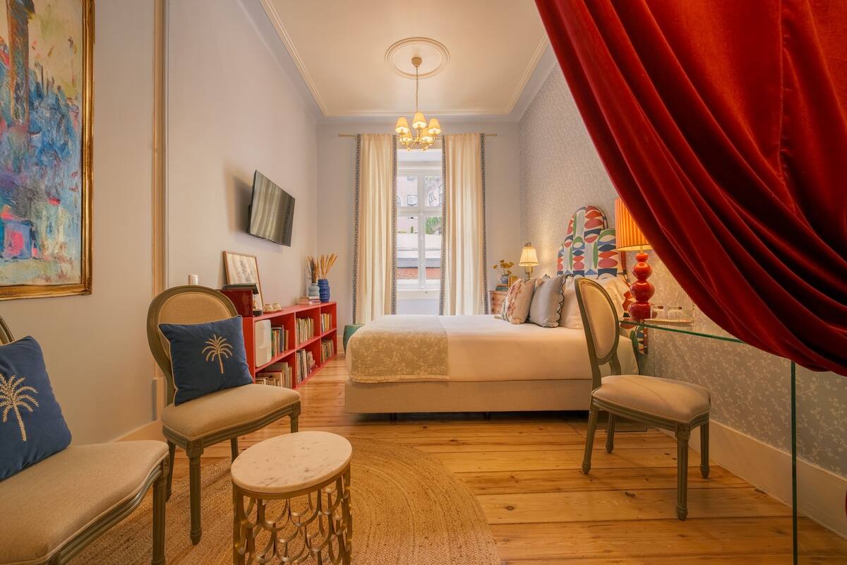 A bedroom with a bed, two chairs, a small table, a TV, a bookshelf, and a window with curtains.
