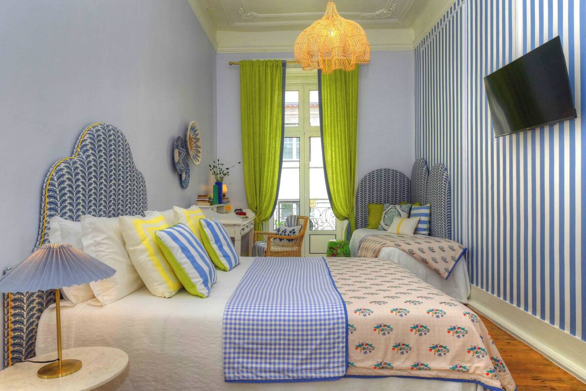 A bedroom with two beds, a desk, a chair, a TV, and a window with curtains.