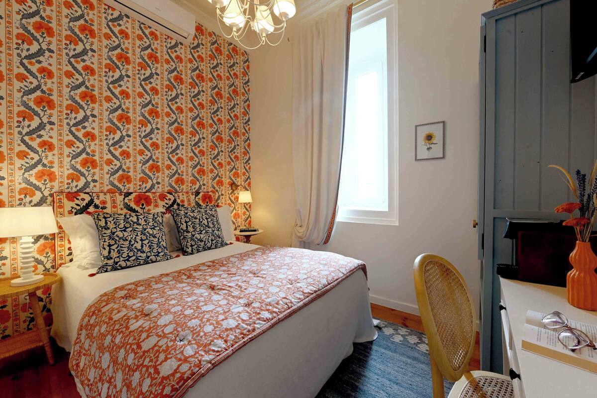A bedroom with a bed, a nightstand, a chair, a desk, and a window with curtains.