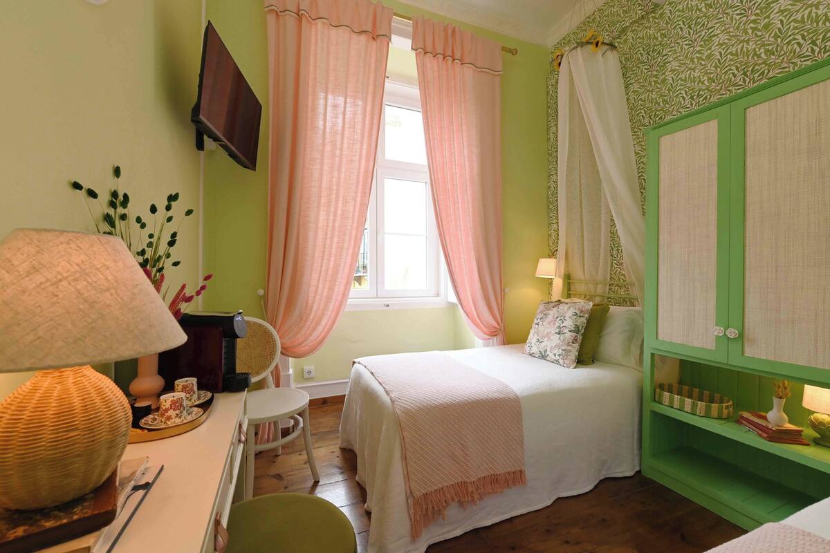 A bedroom with a bed, a desk with a lamp, a chair, a window with curtains, and a green wardrobe.
