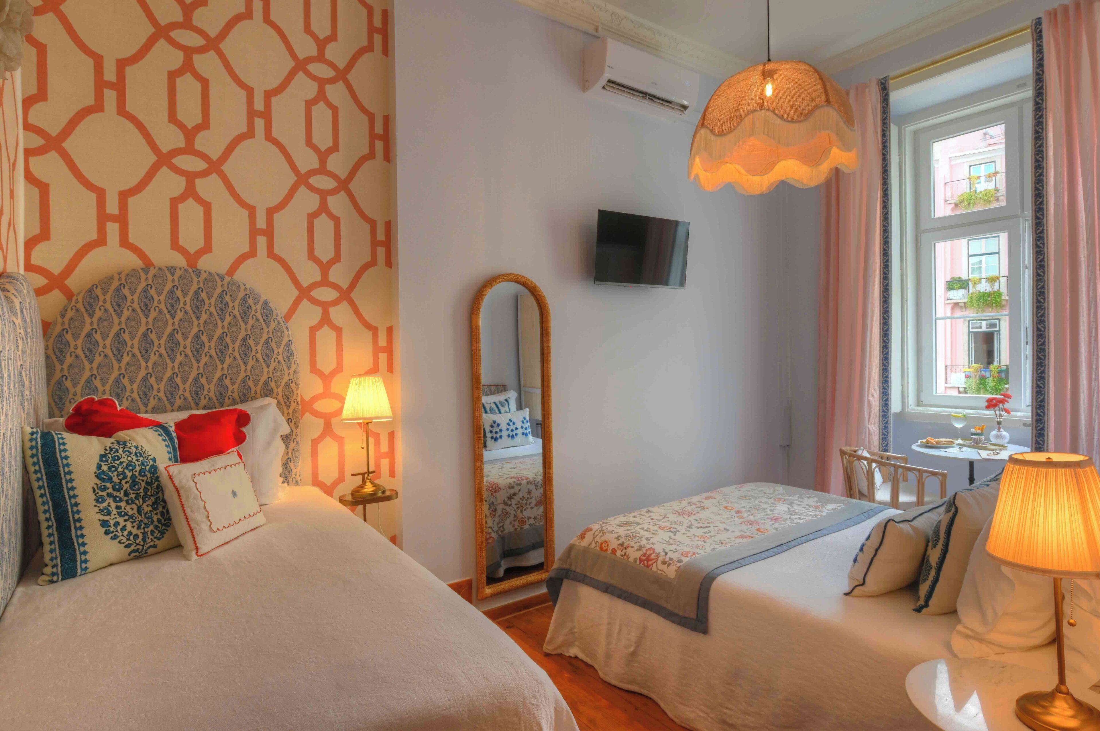 A hotel room with two beds, a TV, a mirror, a window with curtains, and a table with a lamp.