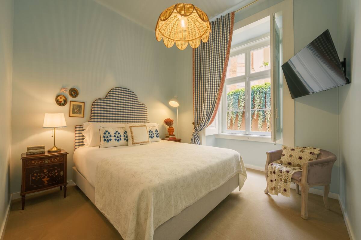 A bedroom with a large bed, a nightstand, a chair, a window with curtains, and a wall-mounted TV.