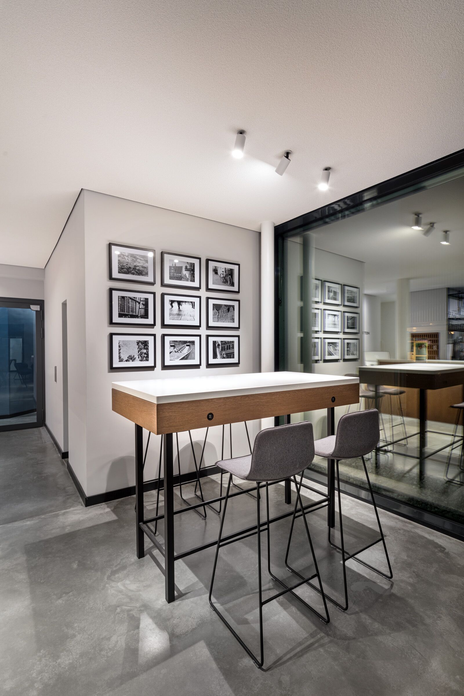 Un comedor moderno con una mesa larga y dos taburetes de bar, una pared con fotografías enmarcadas y un panel de vidrio que separa el comedor de otro espacio.
