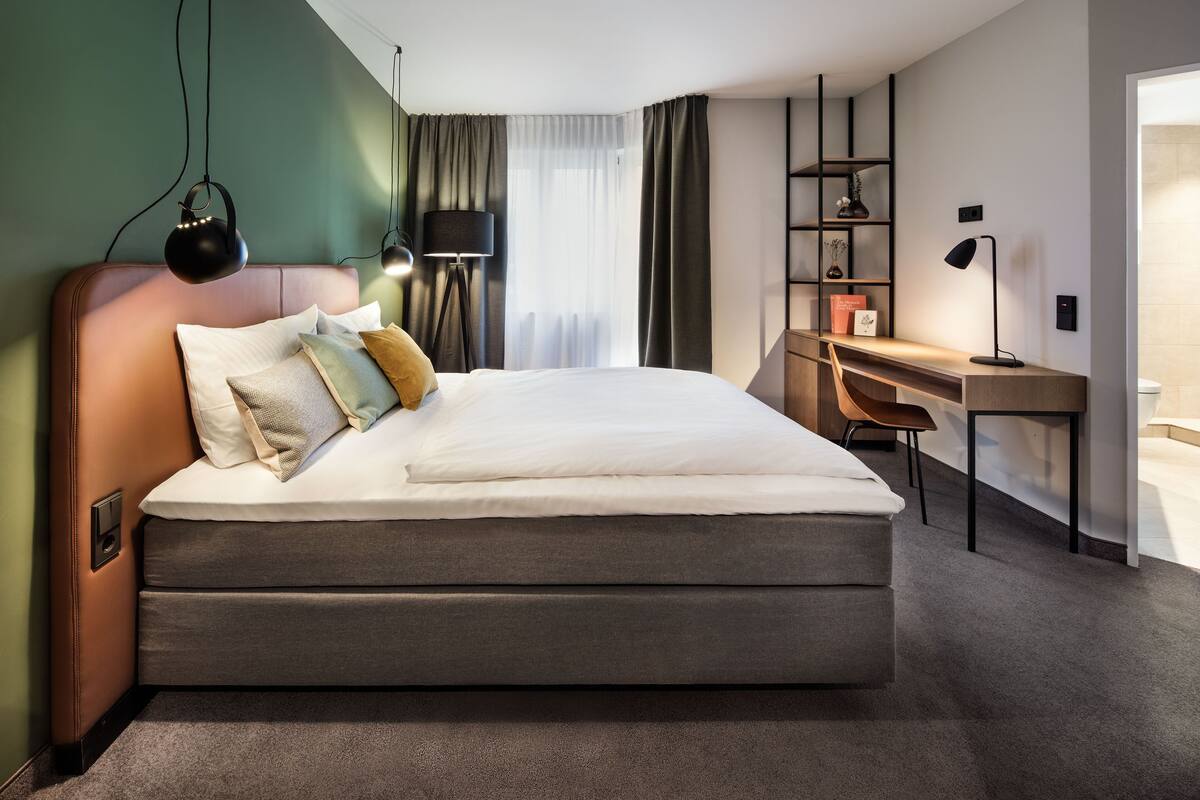 Quarto de hotel moderno com uma cama grande, uma mesa de escritório e uma cadeira. Há uma luminária na mesa e uma prateleira com objetos decorativos.