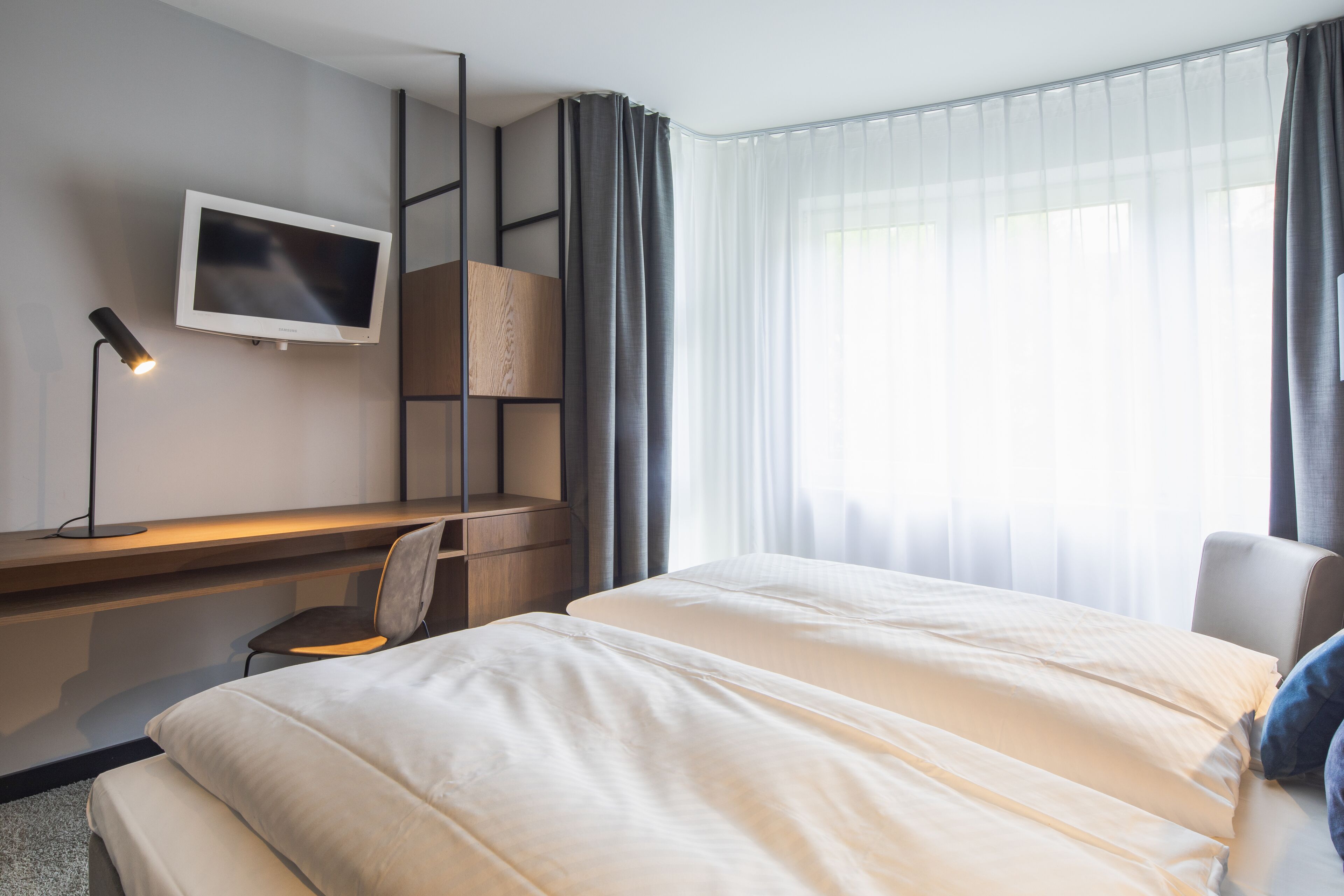 A hotel room with a bed, a desk, a chair, a TV, and a window with curtains.