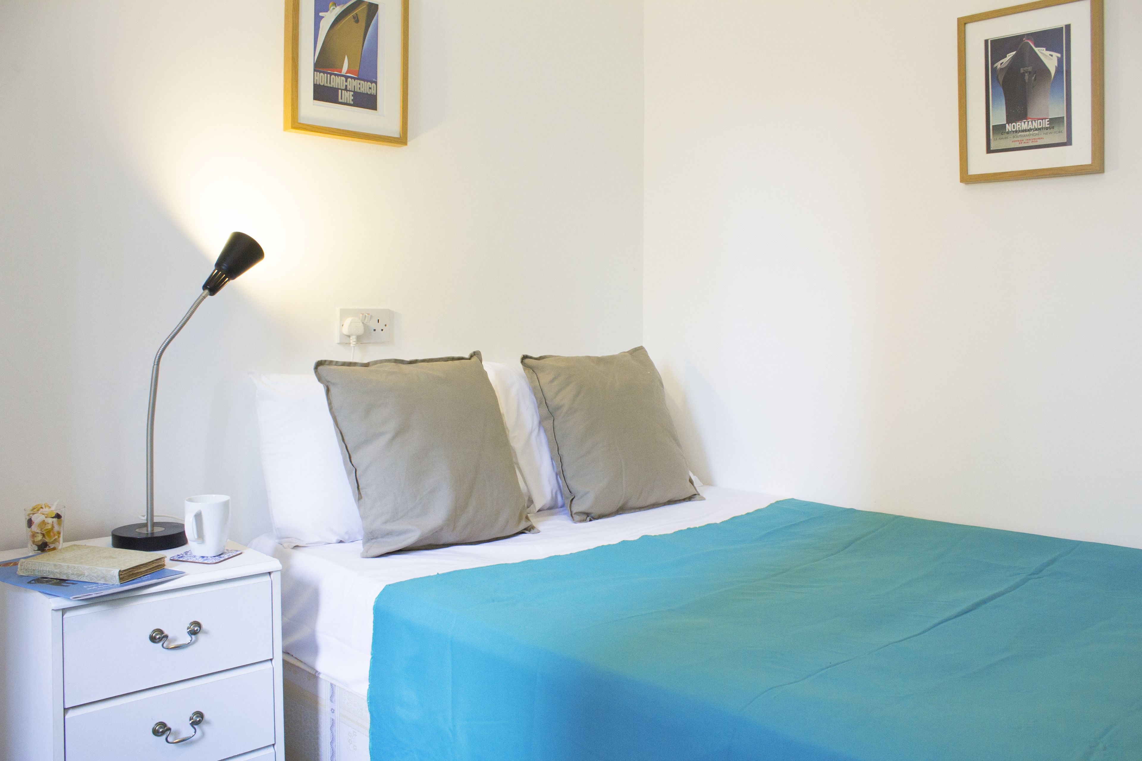 Una cama bien tendida con una colcha azul, dos almohadas, una lámpara de noche y una mesita de noche blanca con cajones.