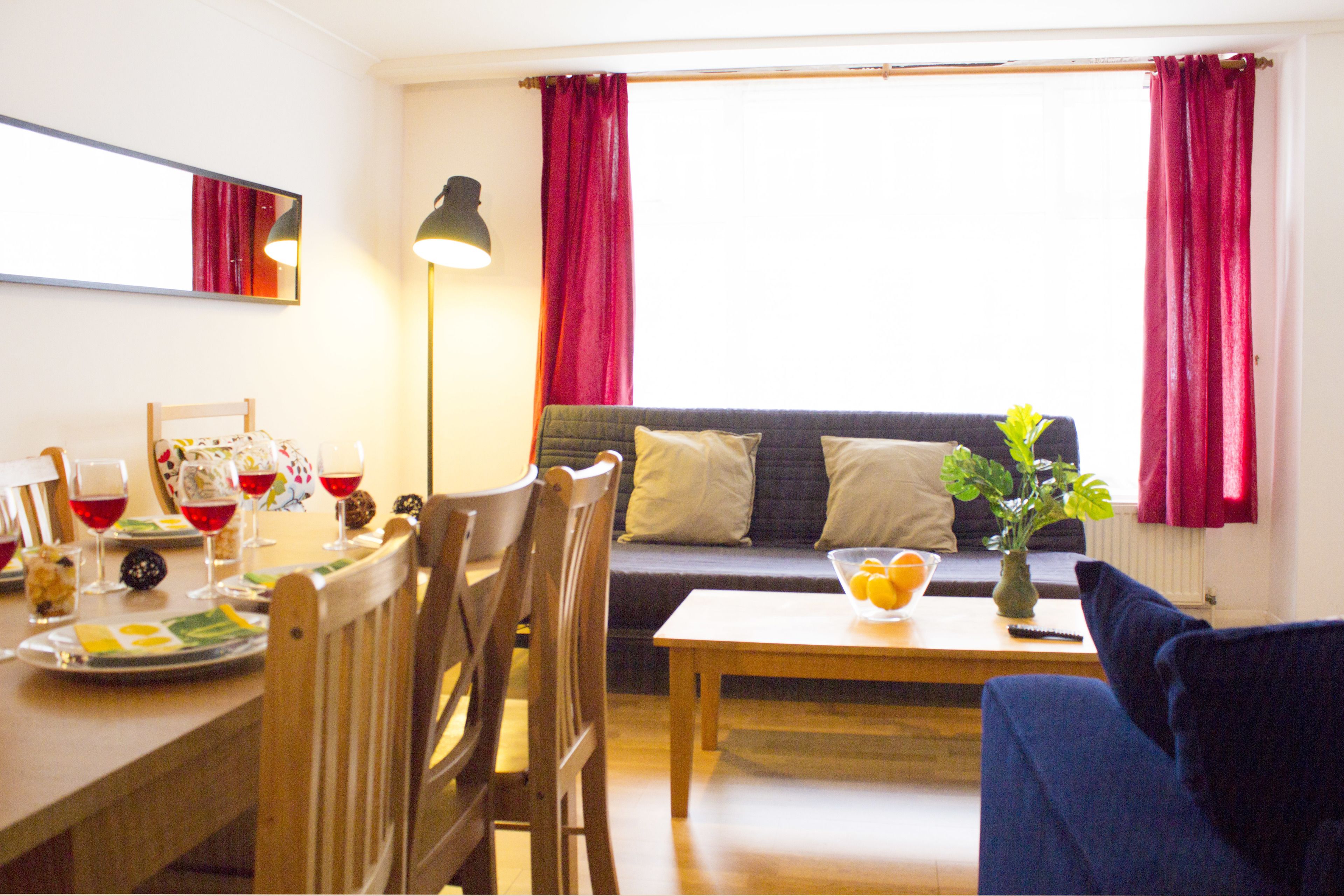 A living room with a sofa, a dining table with chairs, a lamp, and a window with curtains.