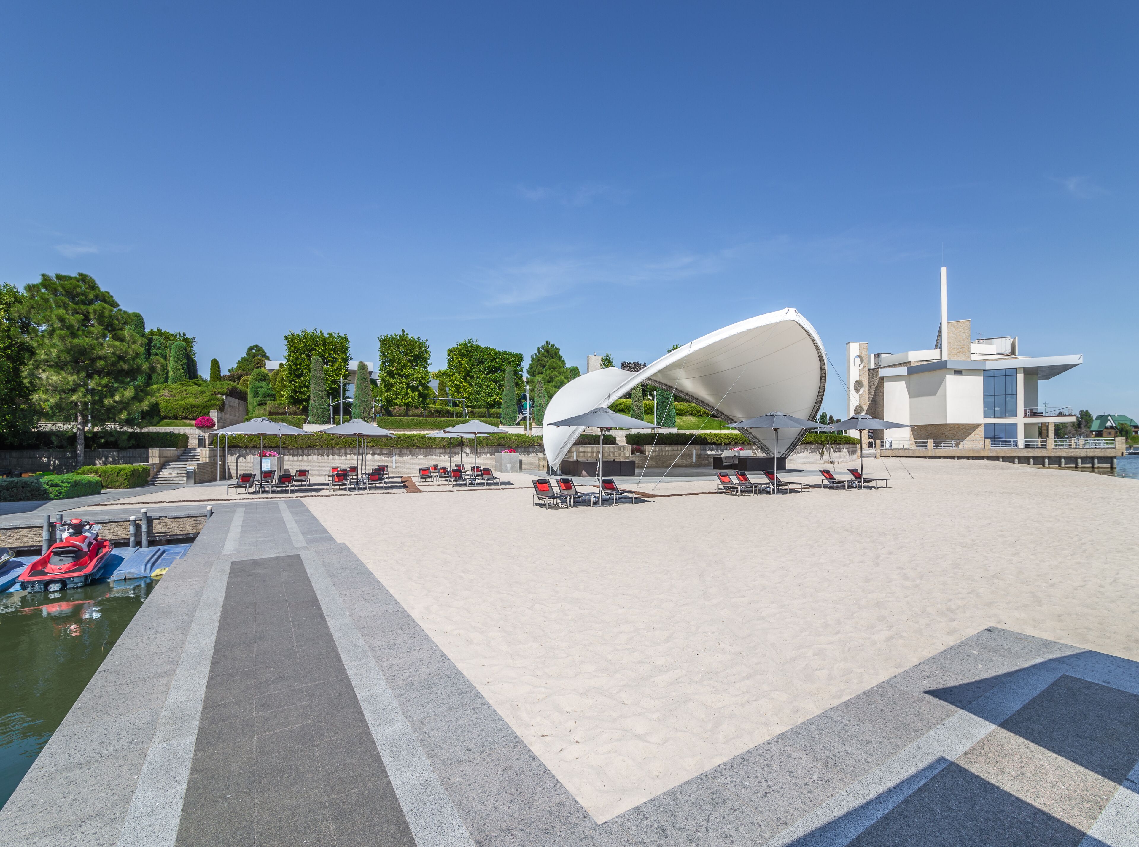 Una estructura arquitectónica moderna con un diseño curvo único, rodeada de una zona arenosa con sillones y sombrillas.