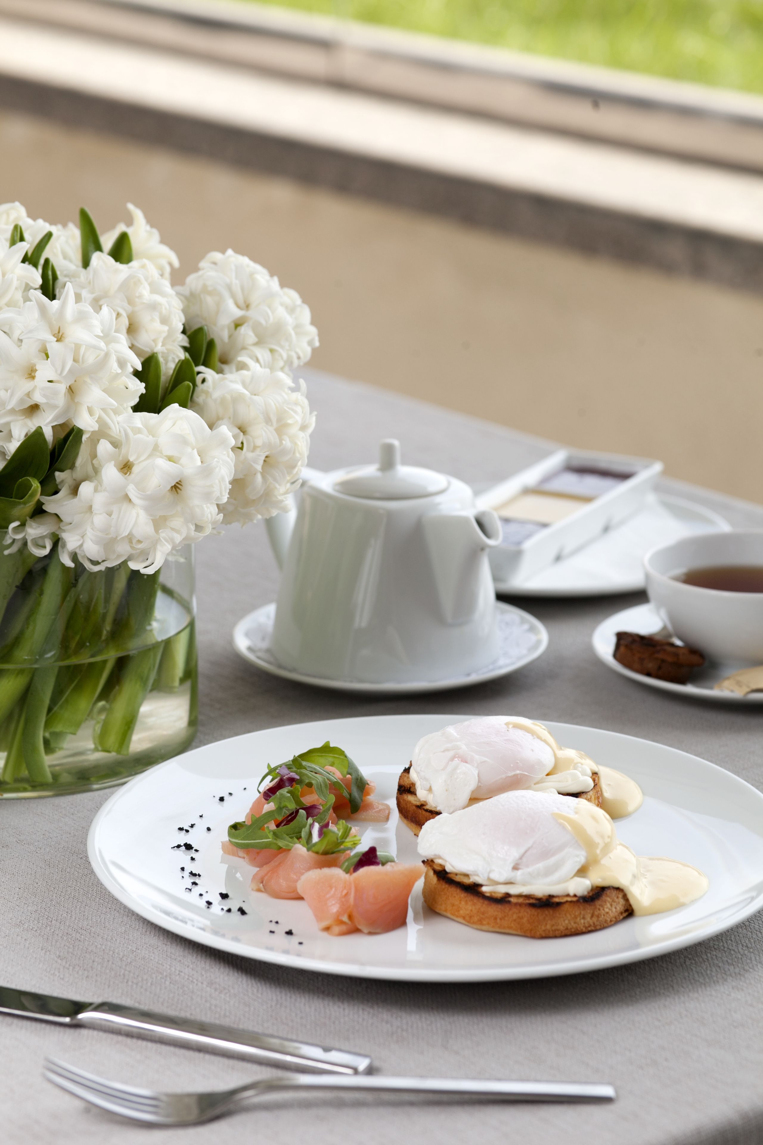 A table with a white teapot, a cup of tea, a plate with food, and a vase of white flowers.
