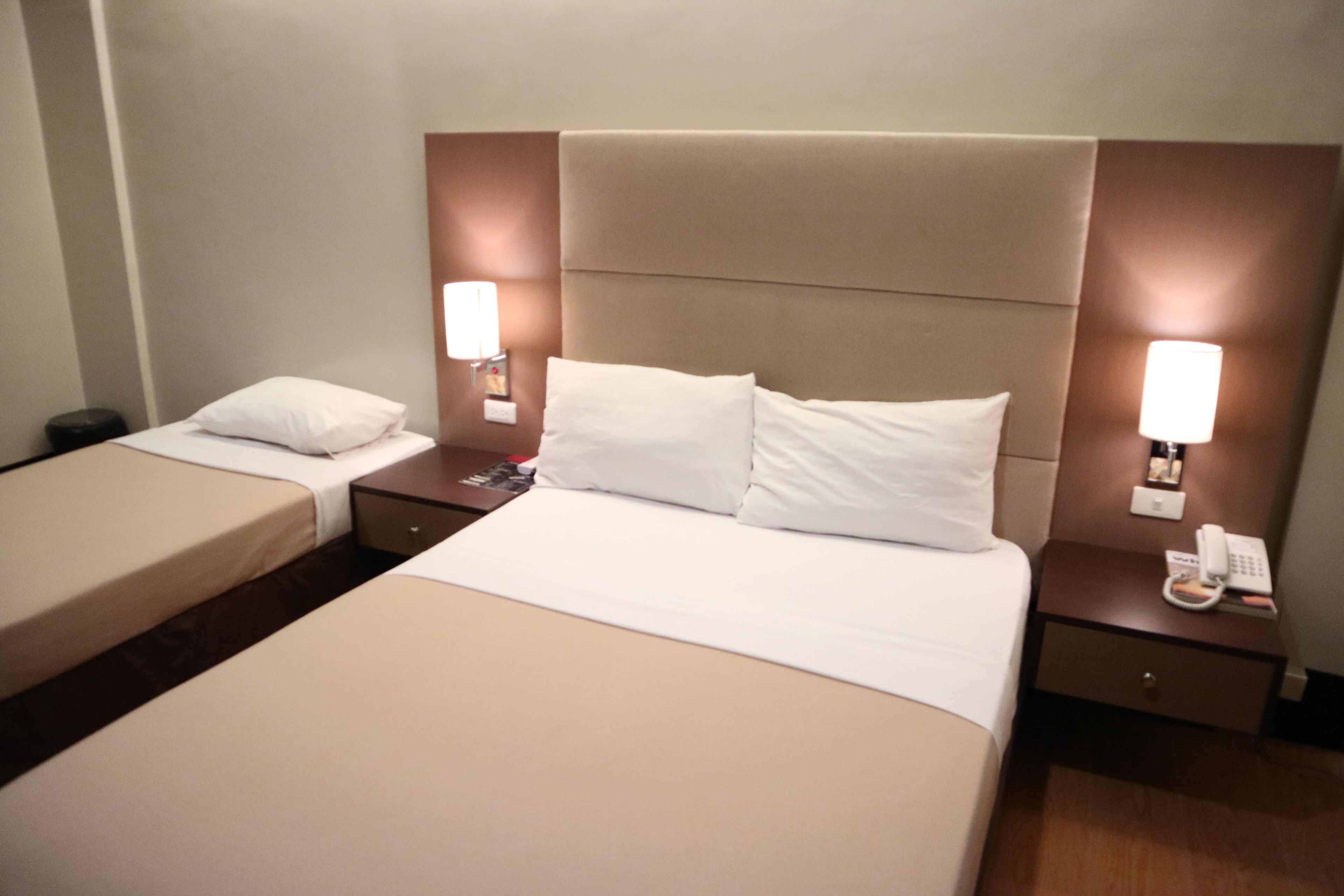 A hotel room with two beds, a nightstand with a phone, and wall-mounted lamps.