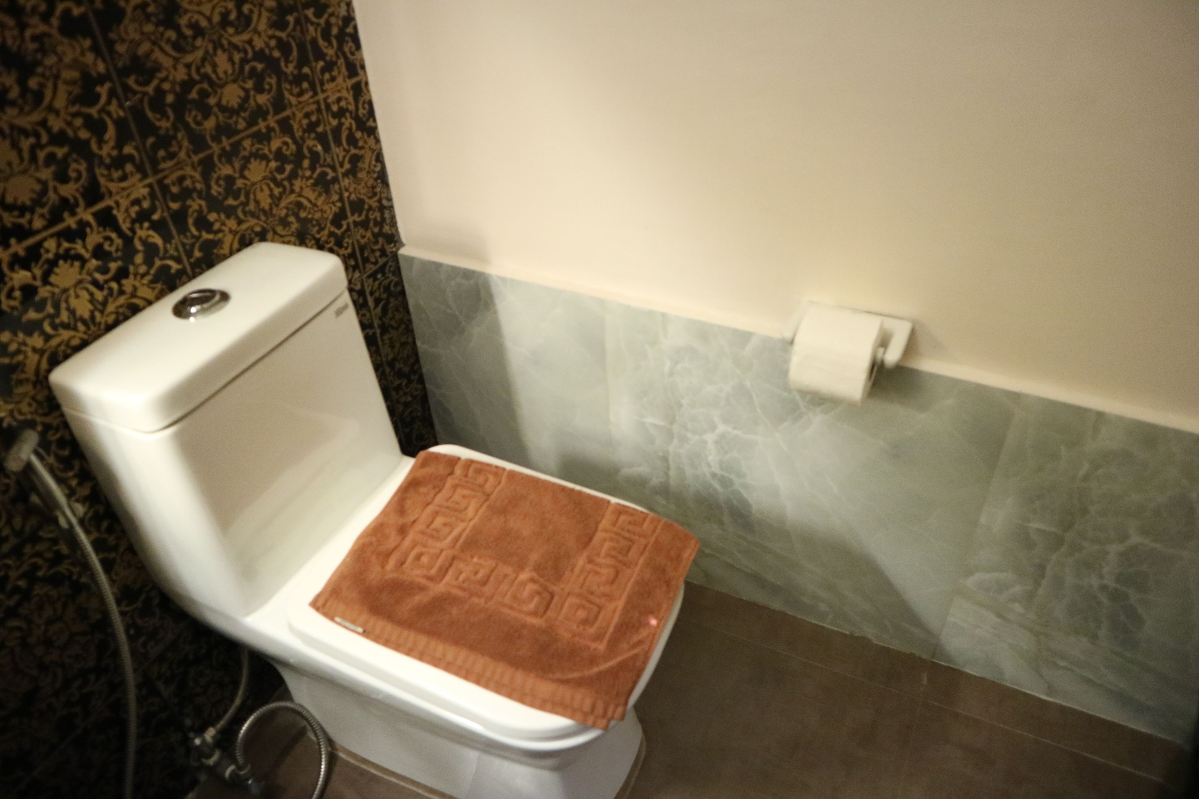 Um sanitário com uma toalha castanha no assento, um rolo de papel higiénico no suporte fixado na parede, e azulejos estampados nas paredes.