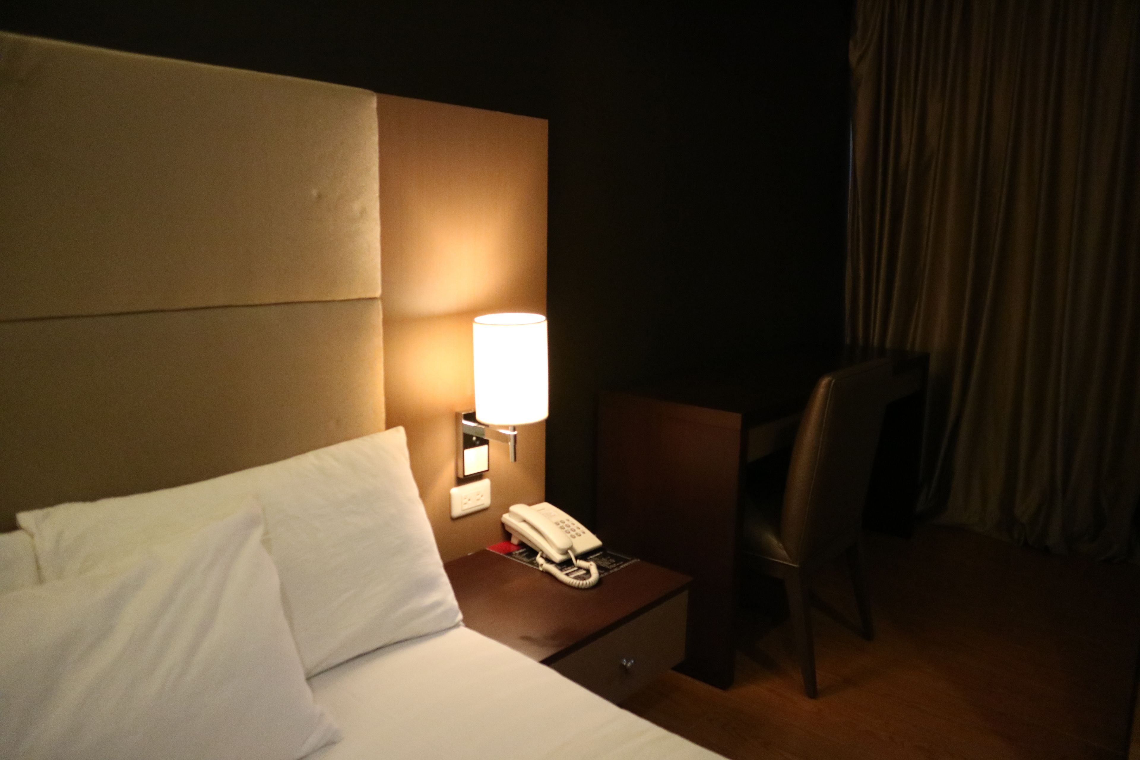 A hotel room with a bed, a bedside lamp, a phone, a chair, and a curtain.