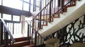Una escalera con escalones de madera y barandillas de metal que conduce a un piso superior. En la zona inferior, hay dos sillones y una pequeña mesa de madera.