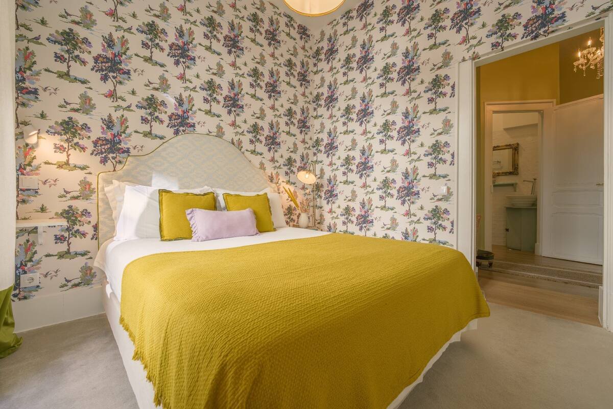 A bedroom with floral wallpaper, a bed with a yellow blanket, and a nightstand with a lamp.