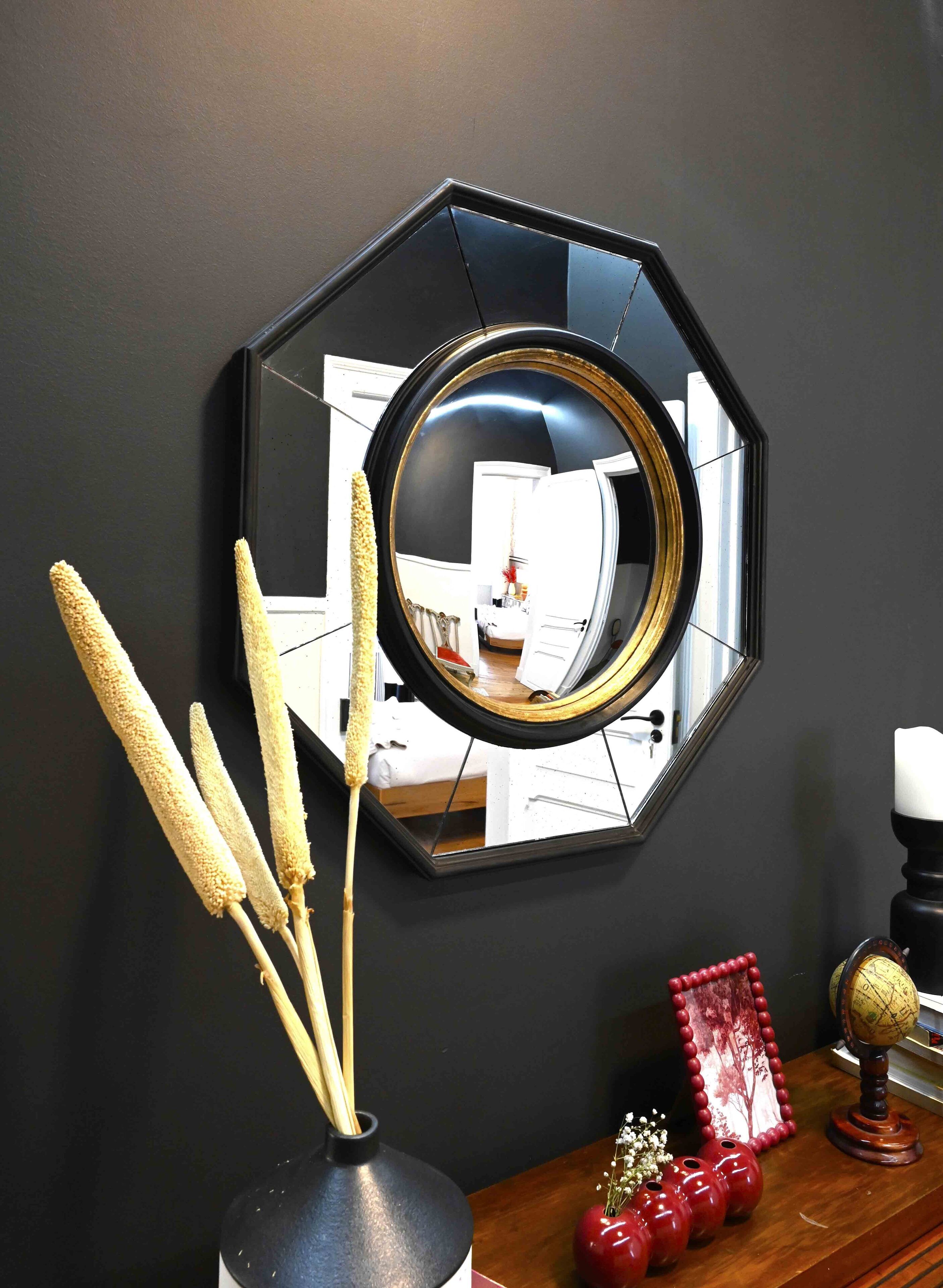 Un miroir hexagonal avec un cadre doré, placé sur un mur sombre, un vase contenant des tiges de maïs séchées, un globe terrestre et un vase rouge orné de fleurs.