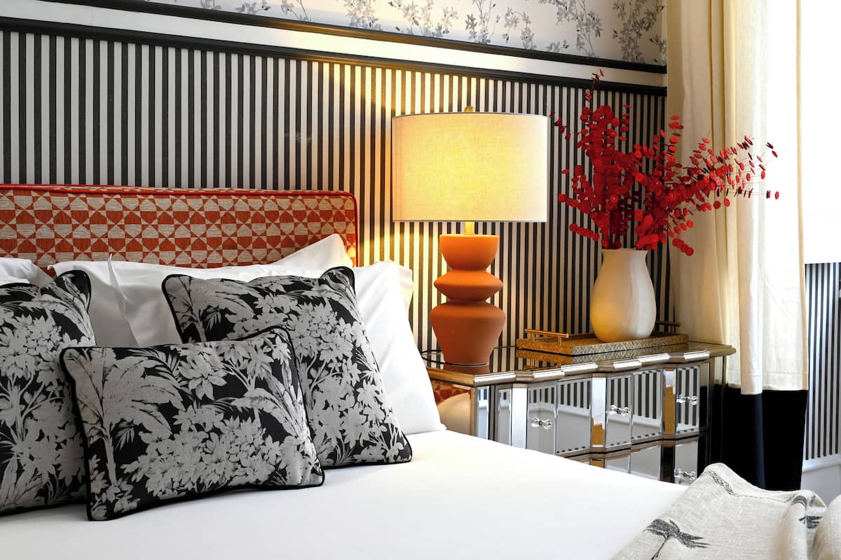 A hotel room with a bed, pillows, a bedside table with a lamp, a vase with red flowers, and a mirror.