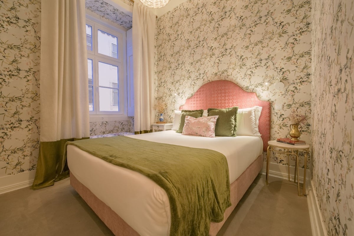 A neatly made bed with a green blanket, a floral wallpapered room, and a window with curtains.