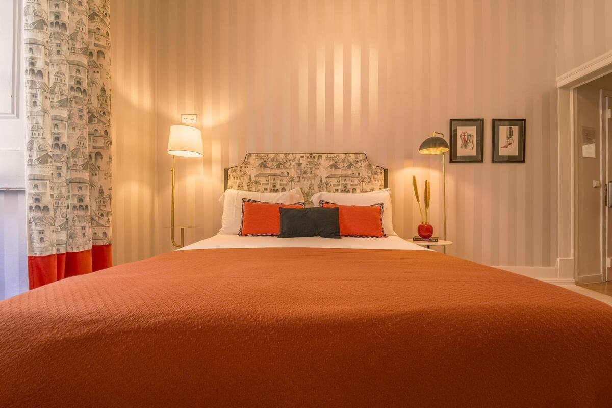 A hotel room with a large bed, striped wallpaper, a nightstand with a lamp, and a door to the right.