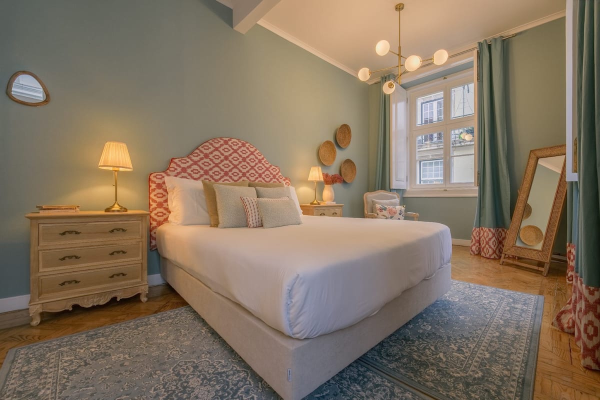 A bedroom with a large bed, two bedside tables, a nightstand, a window with curtains, and a mirror.