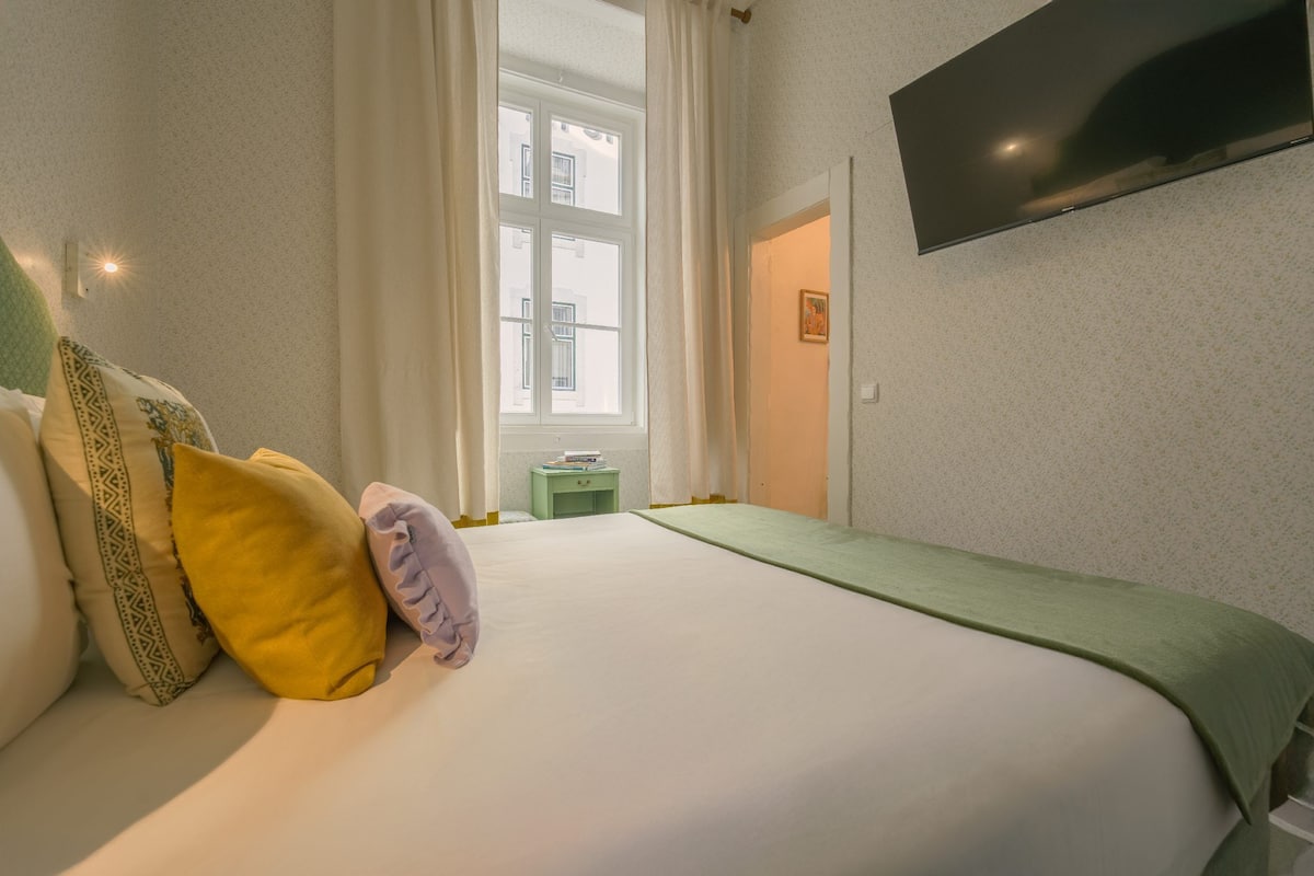 A bedroom with a bed, pillows, a nightstand, a TV, and a window.