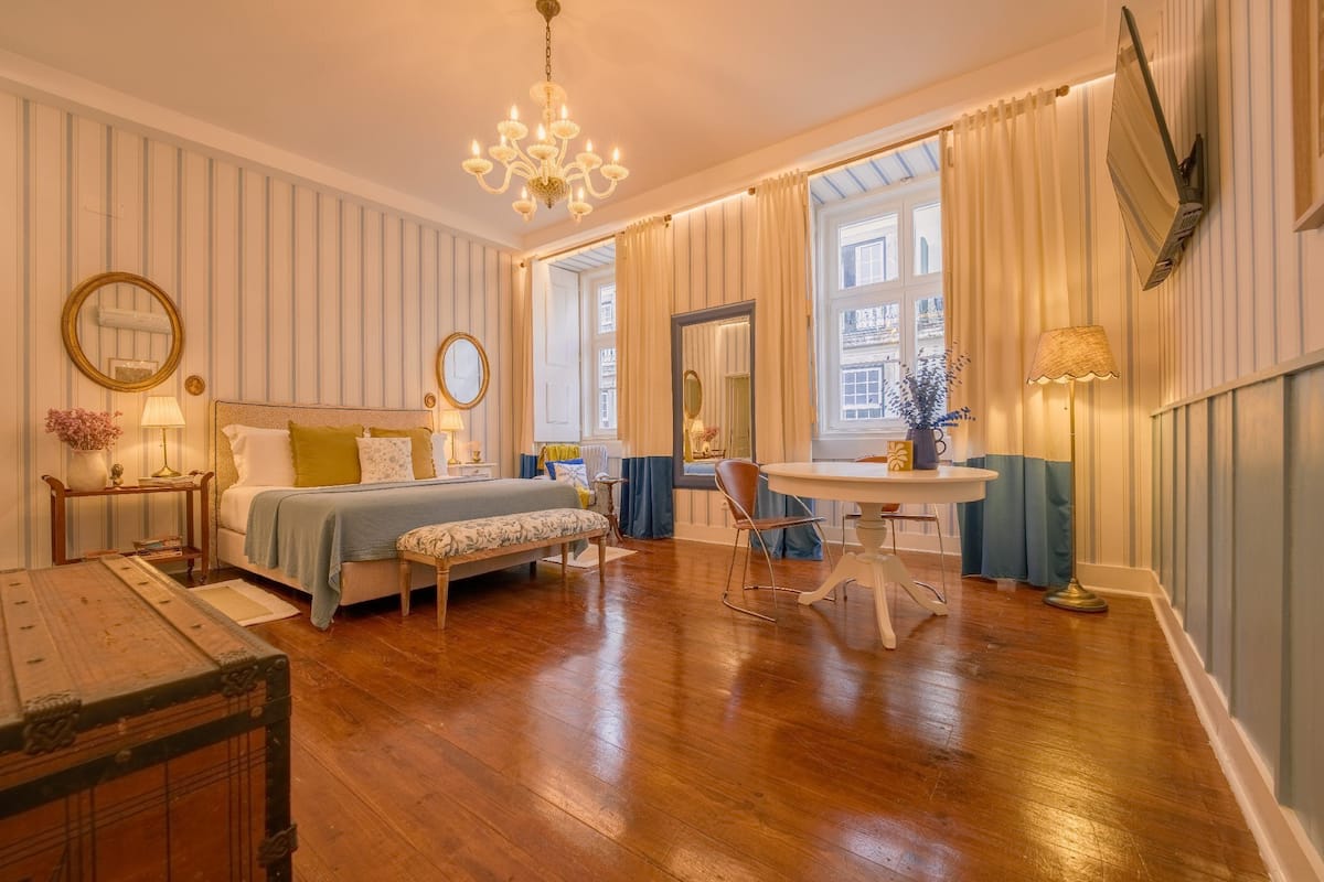 A well-lit bedroom with a wooden floor, a bed with a blue and white bedspread, a bedside table with a lamp, and a large window with curtains.