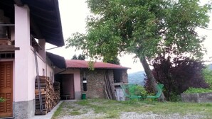 Un edificio rústico con fachada de piedra, puerta de madera y tejado de tejas rojas. Hay una pila de leña afuera y un árbol que da sombra.