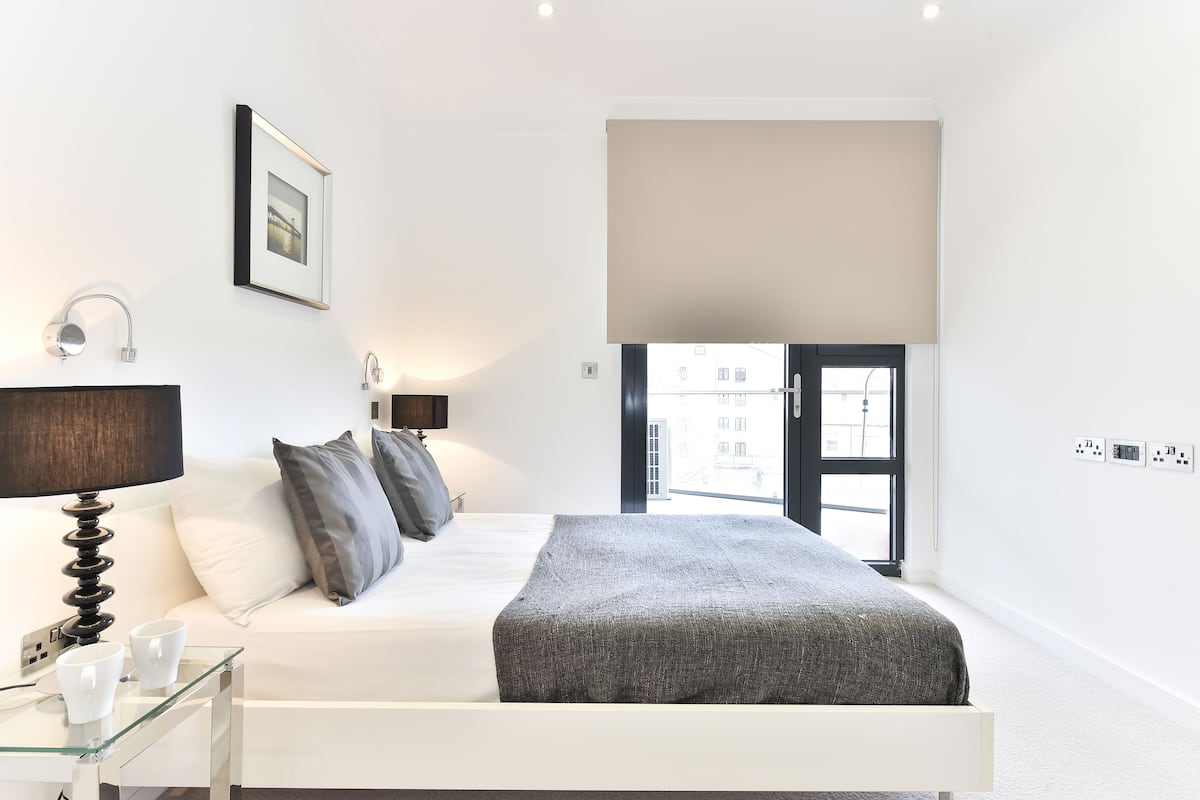 A modern bedroom with a bed, bedside lamps, a glass table with cups, a window with a view, and a large window blind.