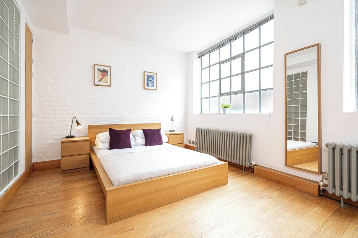 A modern bedroom with a wooden bed, white walls, and a large window.