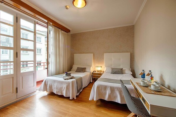 A hotel room with two beds, a small table with cups and bottles, a chair, and a view of buildings through the window. - Hostal Zahara (Ferrol)