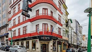 A building with a sign reading "Hostal zafora" on the ground floor, featuring a red and white facade, multiple windows, and a cobblestone street in front. - Hostal Zahara (Ferrol)