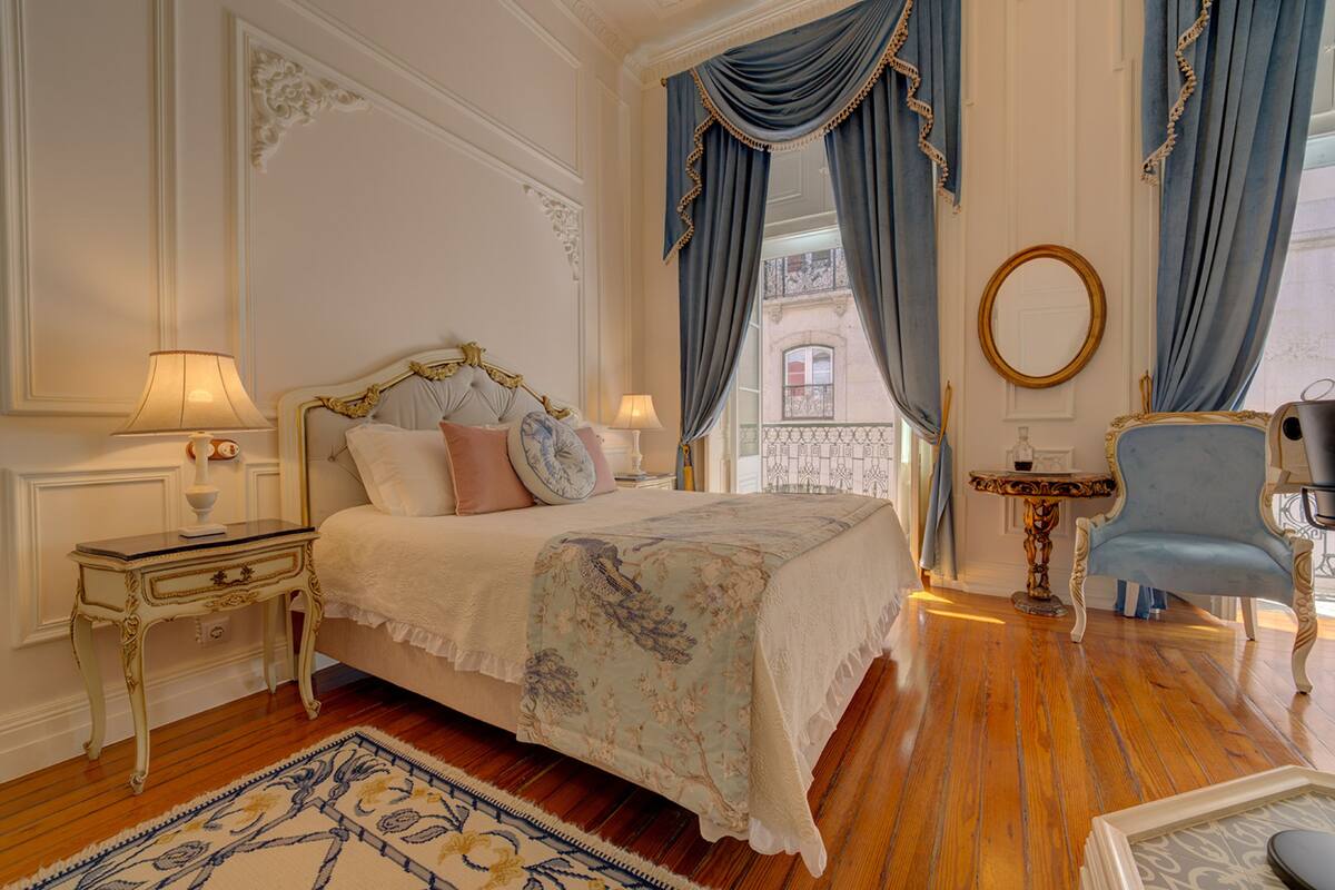 Een klassieke slaapkamer met een hemelbed, een tafeltje met een lamp, een stoel en uitzicht op een balkon.