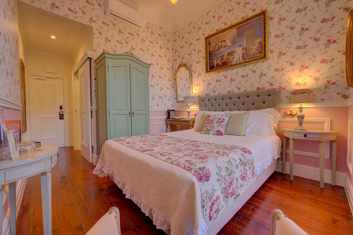 Een slaapkamer met bloemenbehang, een groot bed met een gestuukt hoofdeinde, een dressoir, een spiegel en een nachtkastje met lampen.