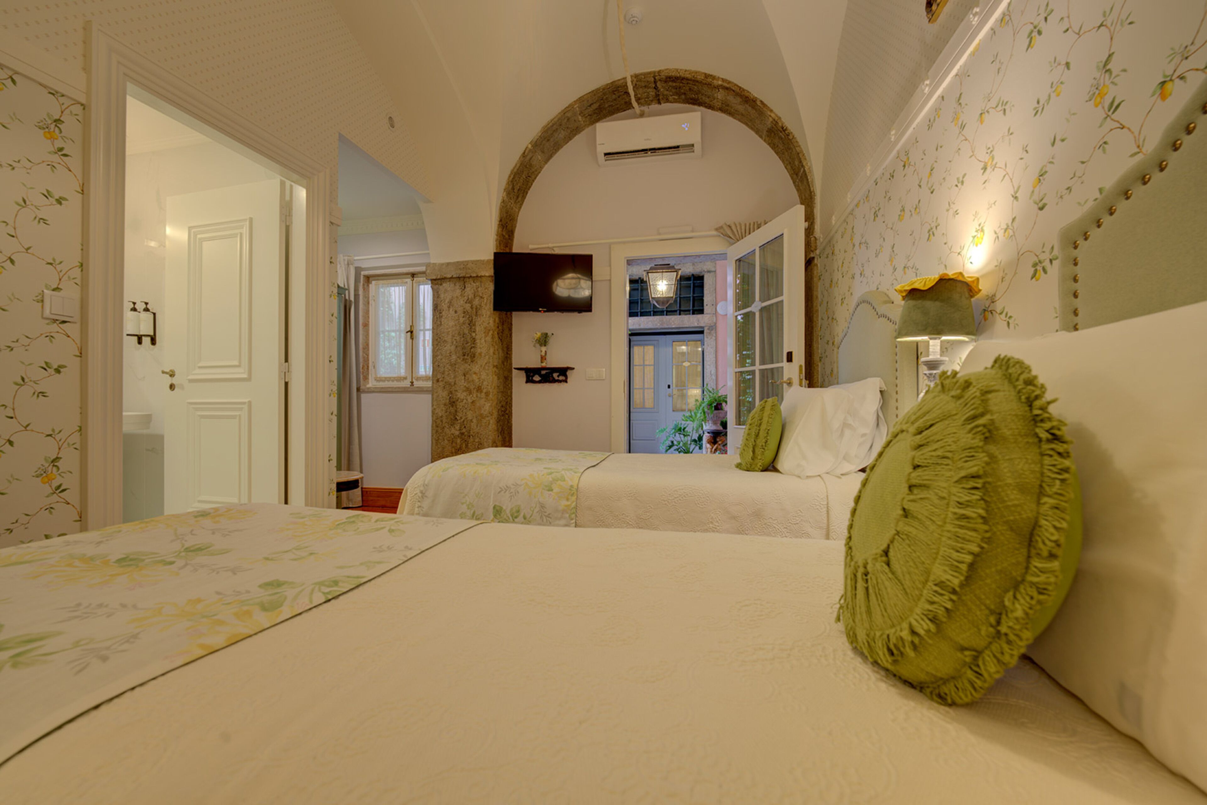A bedroom with a large bed, a smaller bed, and a sofa. There is a television on the wall and a window with curtains.