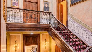 A grand staircase with ornate railings and a red carpet, leading to a second-floor landing with a wooden door and a framed portrait. - Palácio das Especiarias (Lisbon)