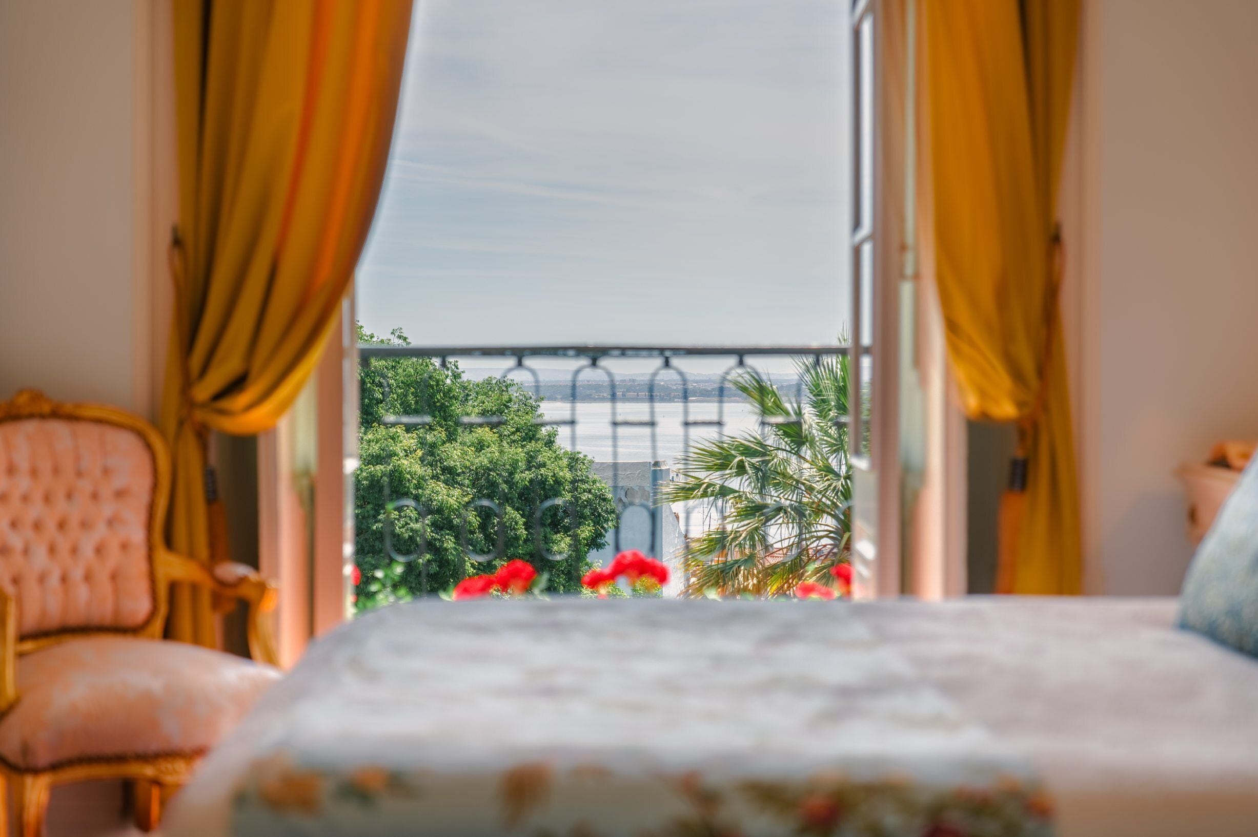 A room with a bed, yellow curtains, a balcony with a view of greenery and a bridge.