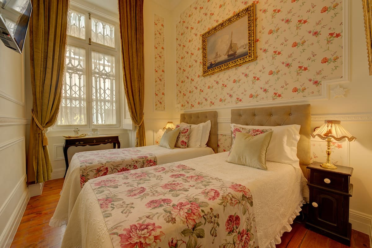 Een hotelkamer met twee bedden, bloemenbehang, een schilderij aan de muur en een raam met gordijnen.
