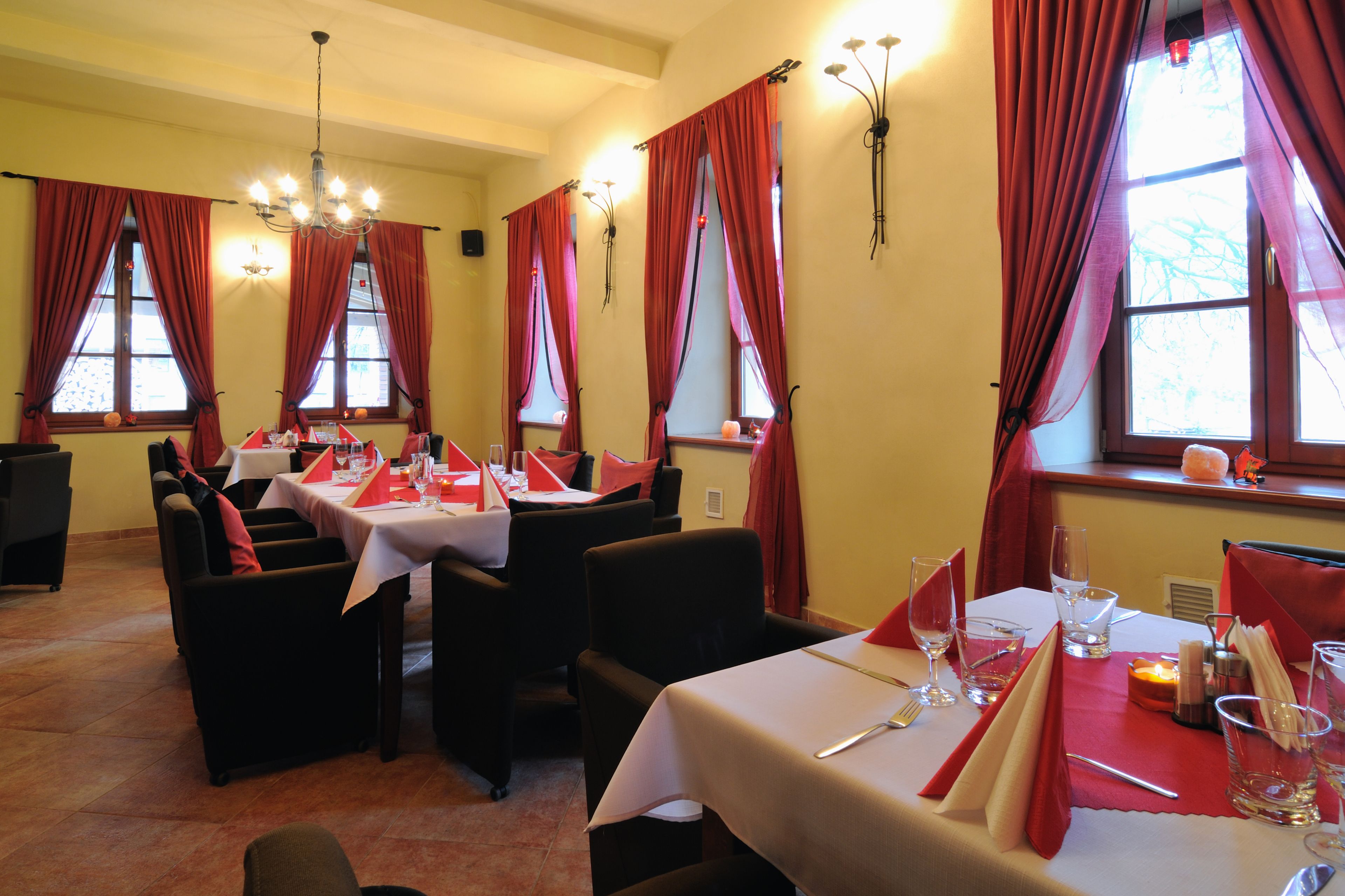 A dining area with tables set for two, red curtains, and a chandelier.
